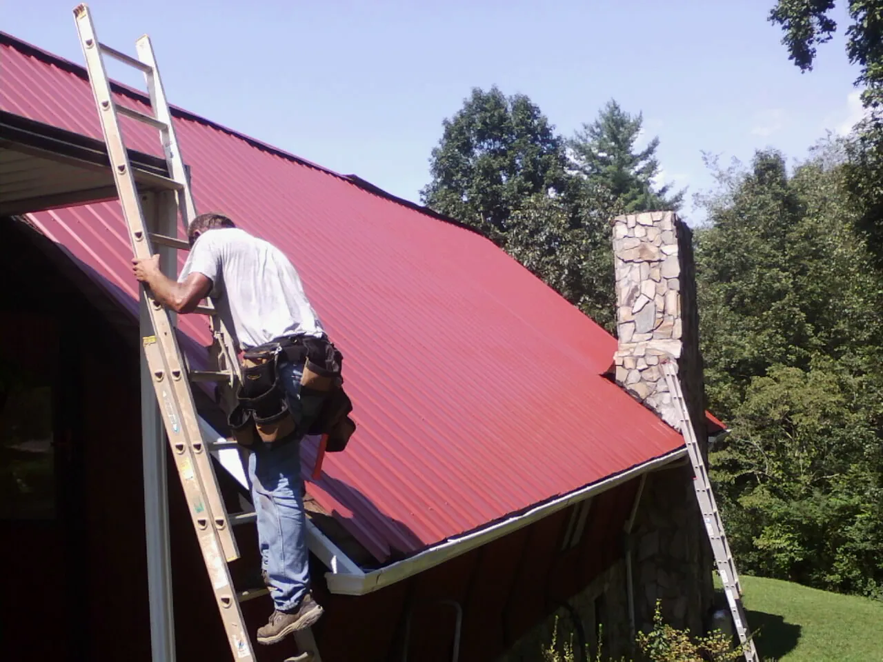 Roofer on a ladder beside a red metal roof and stone chimney — Soffit & Fascia Repair by Roofing Brothers in Woodland Park